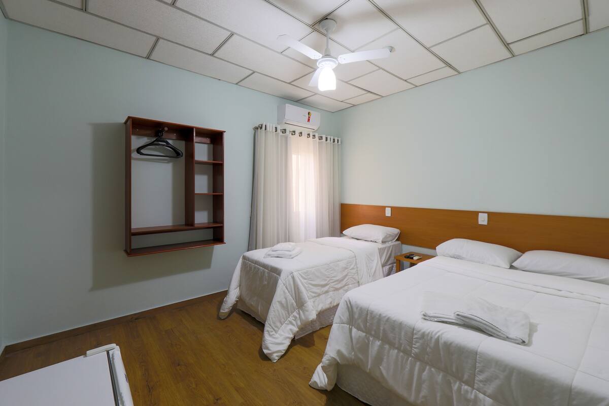 A hotel room with two single beds, a wooden shelf with a hanger, a ceiling fan, and an air conditioning unit.