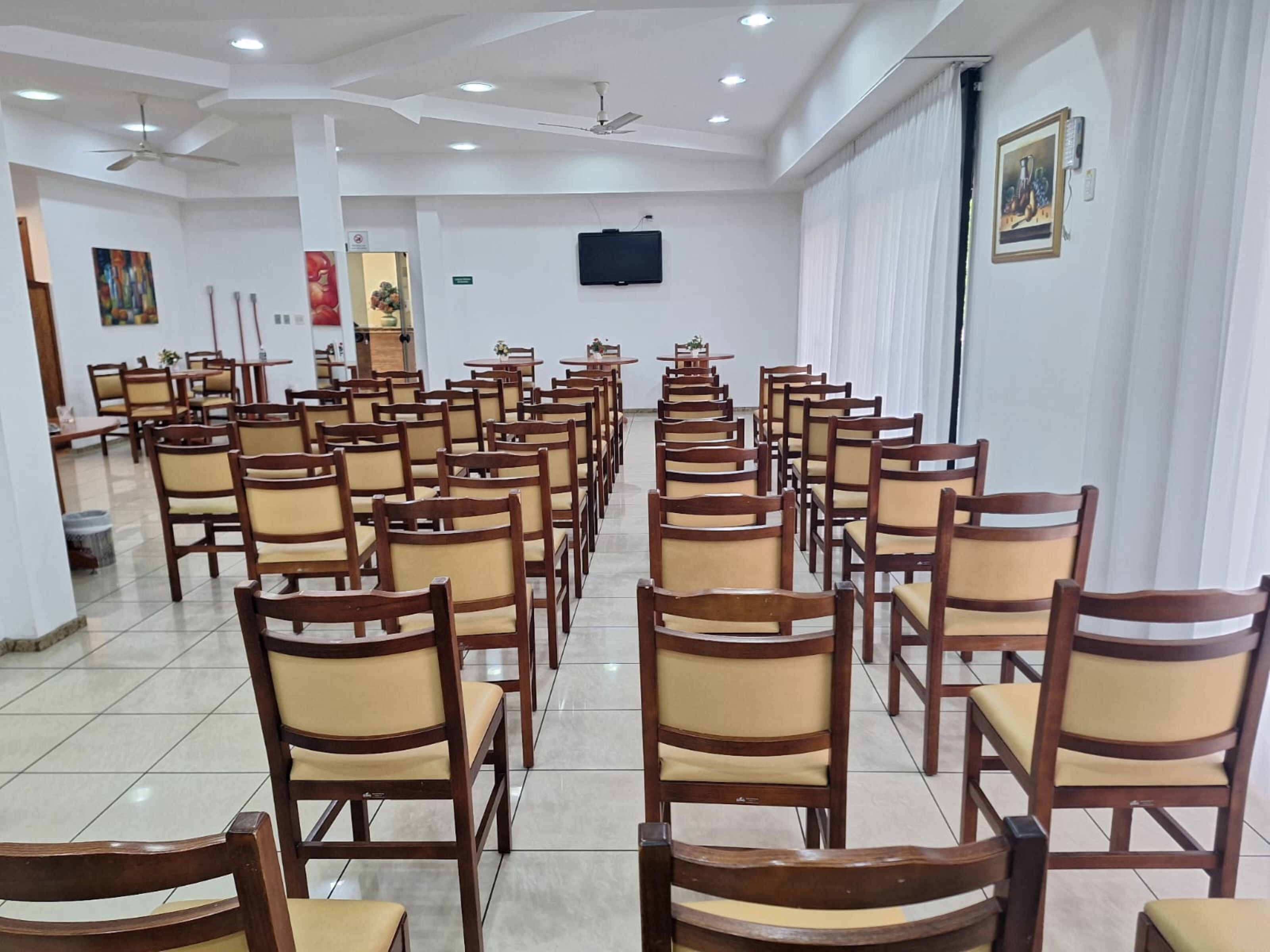 A conference room with rows of wooden chairs and tables, white walls, ceiling fans, and a mounted television.