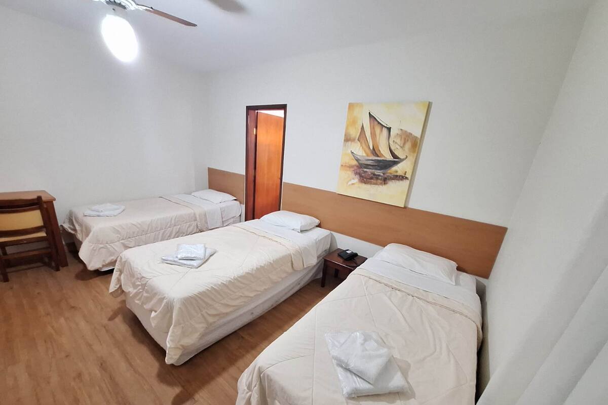 A hotel room with three single beds, a wooden desk, a chair, and a painting on the wall.