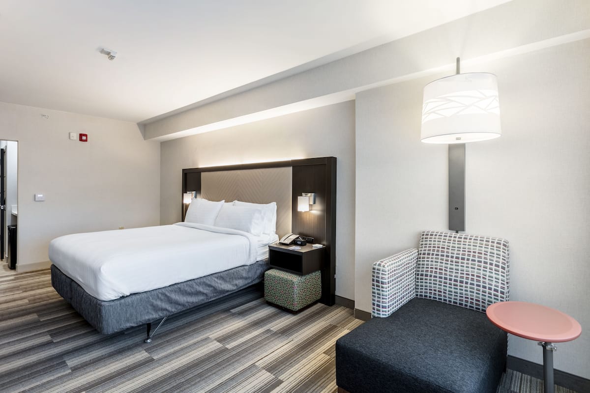 A modern hotel room with a large bed, a bedside table with a lamp, a small seating area, and a wall-mounted light fixture.