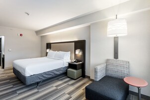 Una habitación de hotel moderna con una cama grande, una mesita de noche con lámpara, una pequeña zona de estar y un aplique de pared.