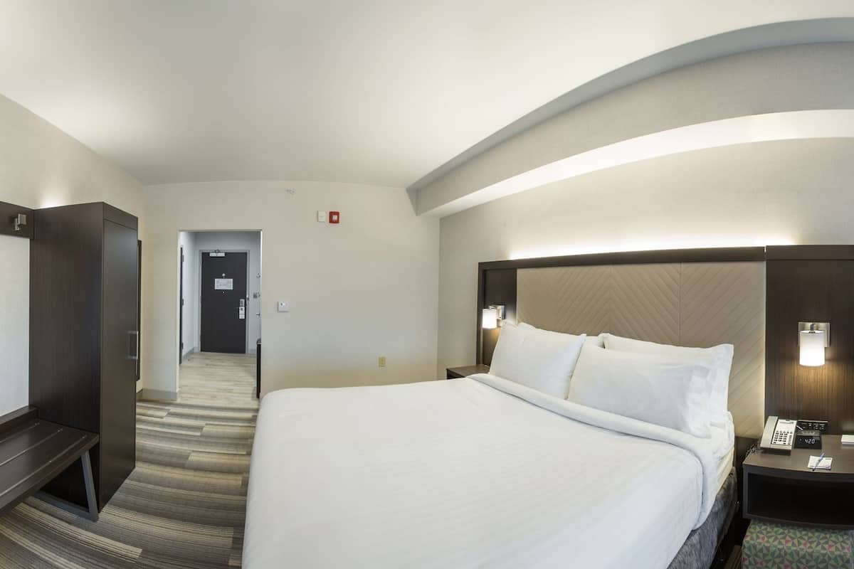 A hotel room with a large bed, a desk with a flat-screen TV, a chair, a small table, and a window with curtains.