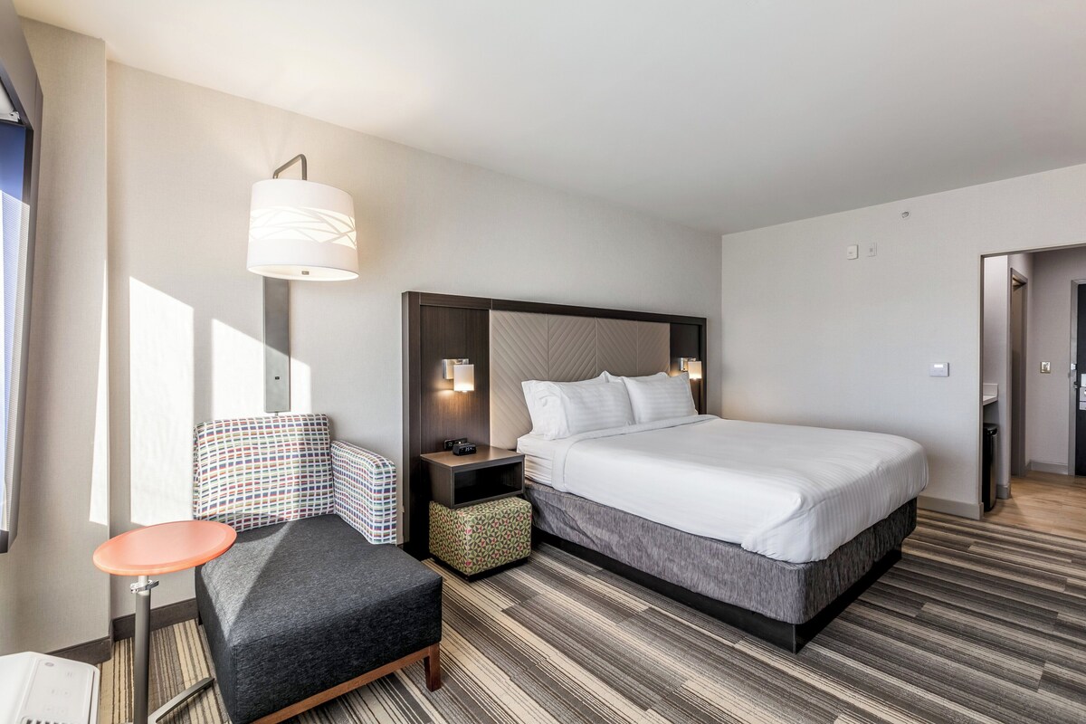 A hotel room with a bed, bedside table, lamp, chair, and a small table.