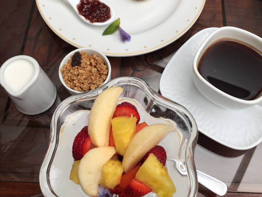 A breakfast setting with a plate of rolls, jam, and a small bowl of granola. There's a glass bowl of fruit and a cup of coffee on the table.