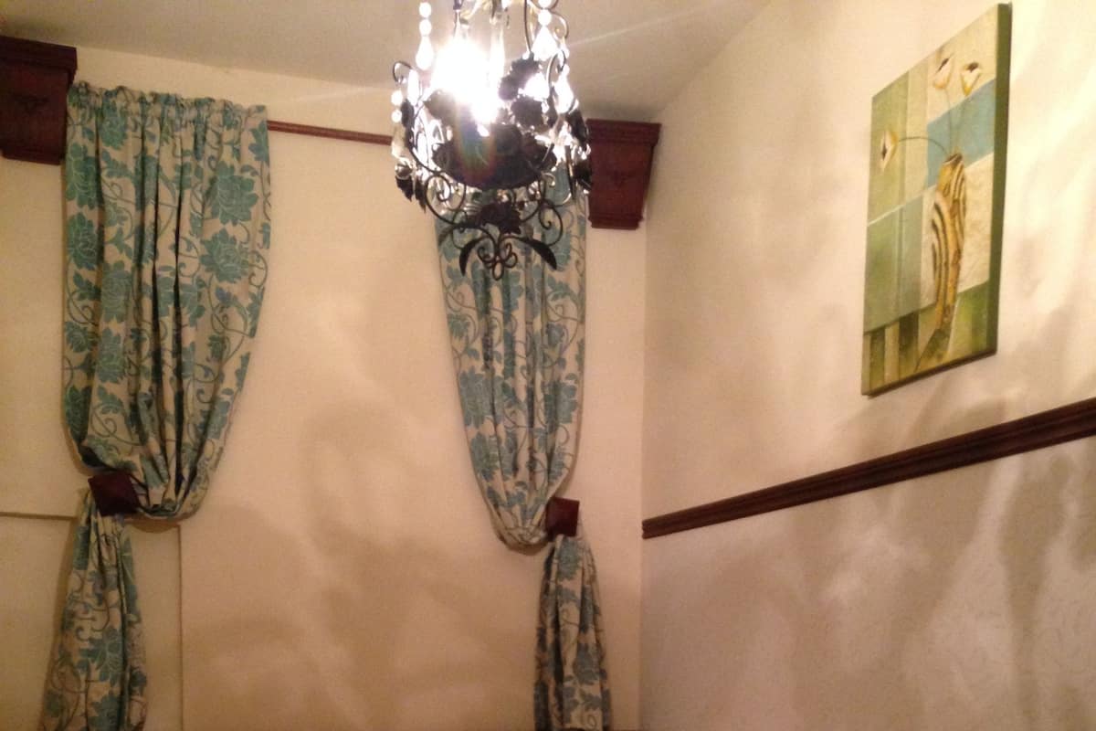 Une chambre à coucher avec un lit, des rideaux, une table de chevet, une lampe, une chaise et un tableau accroché au mur.