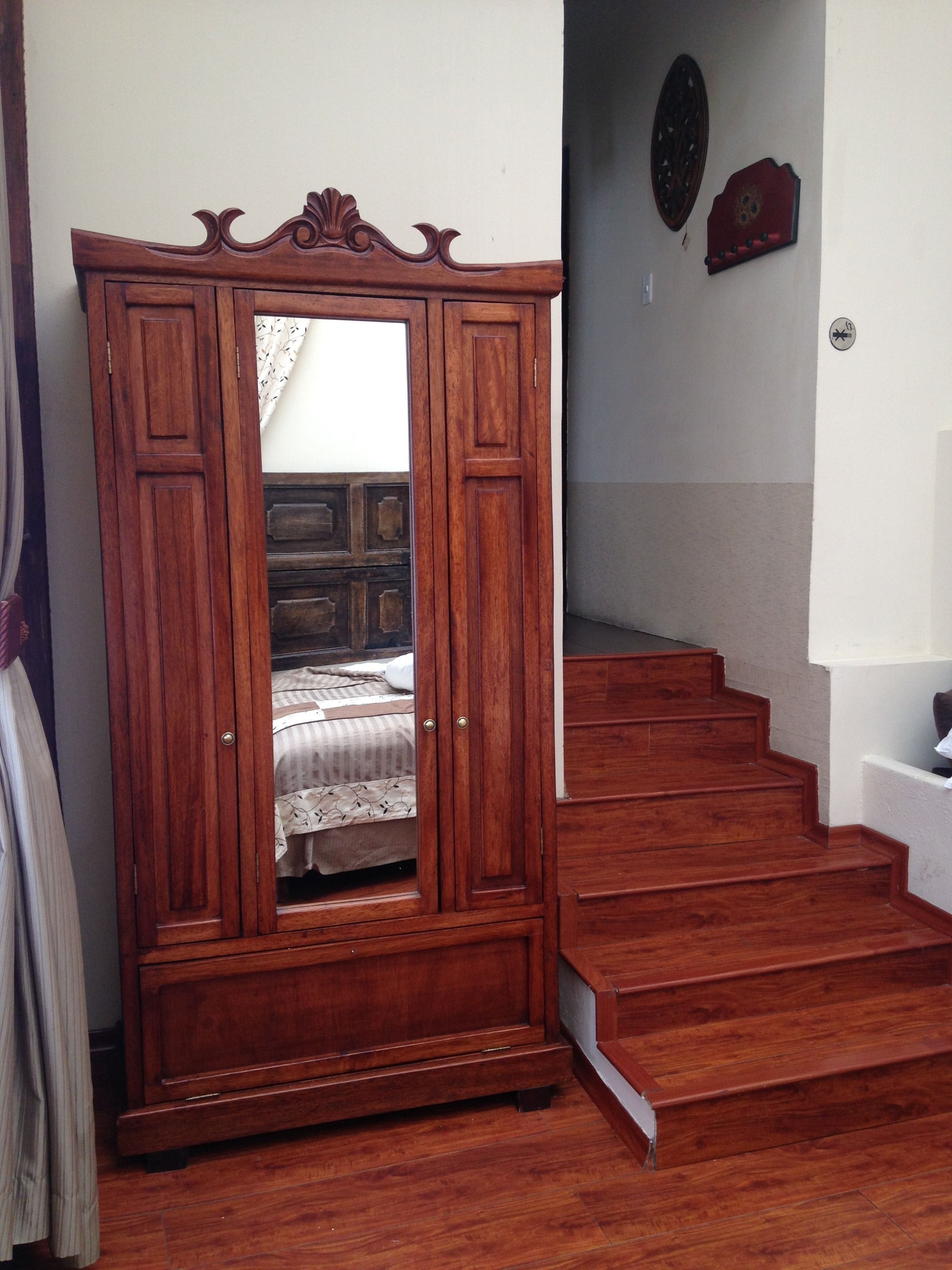 A wooden wardrobe with a mirror, a bed with a patterned bedspread, and a staircase leading to an upper level.