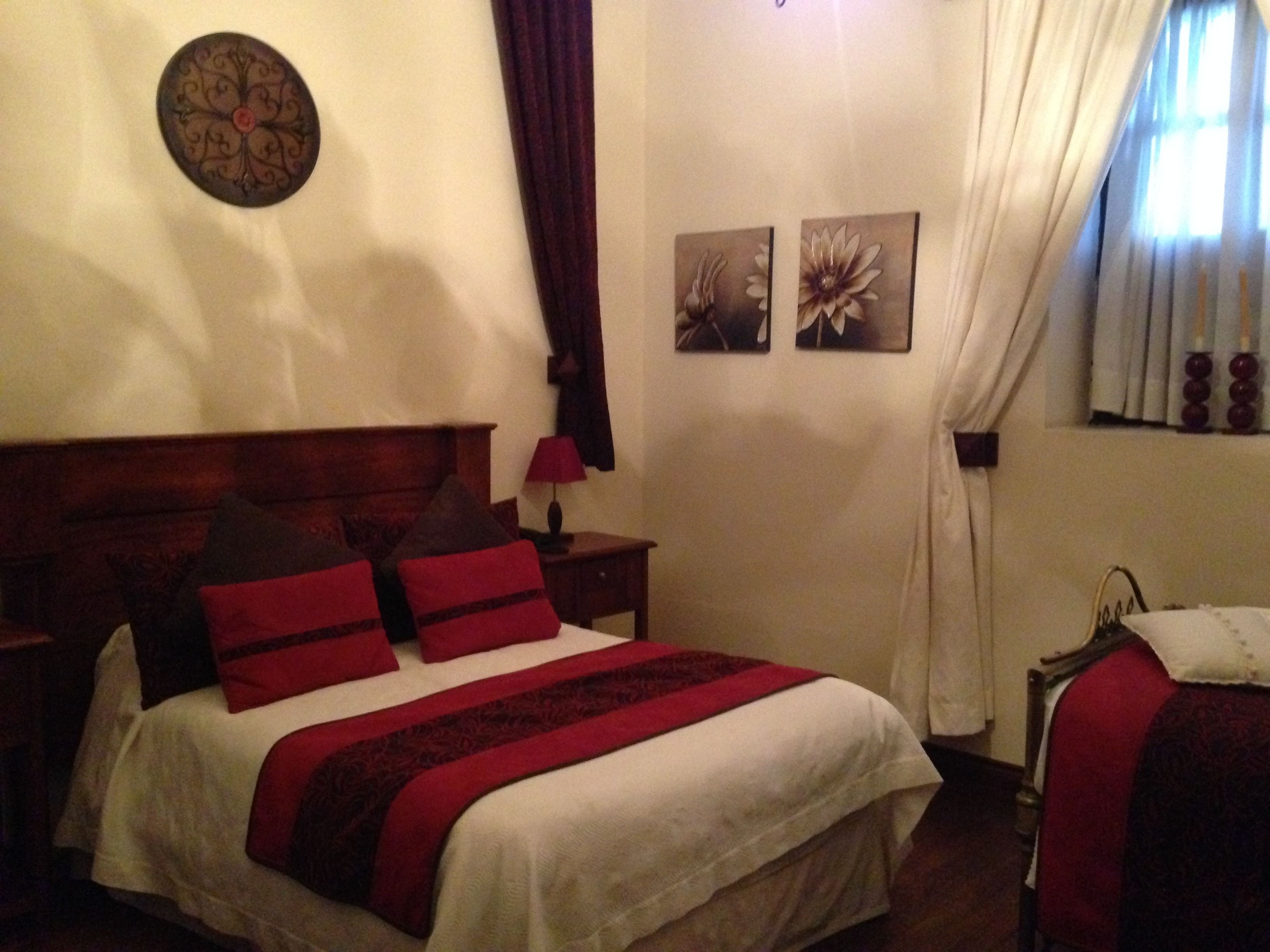 Una cama bien hecha con ropa de cama roja y blanca, un cabecero de madera y un reloj de pared. Hay dos cuadros en la pared y una ventana con cortinas.