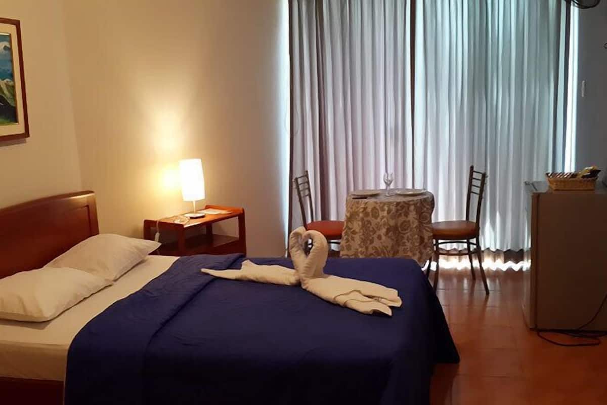 Quarto de hotel com cama, escrivaninha, cadeira, mesa e um frigorífico.