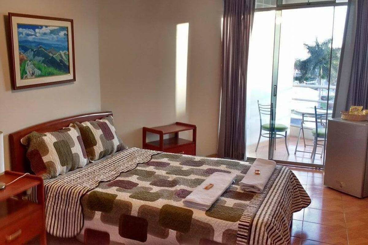 Quarto de hotel com cama, mesa de cabeceira, luminária, um quadro na parede e vista para o exterior.
