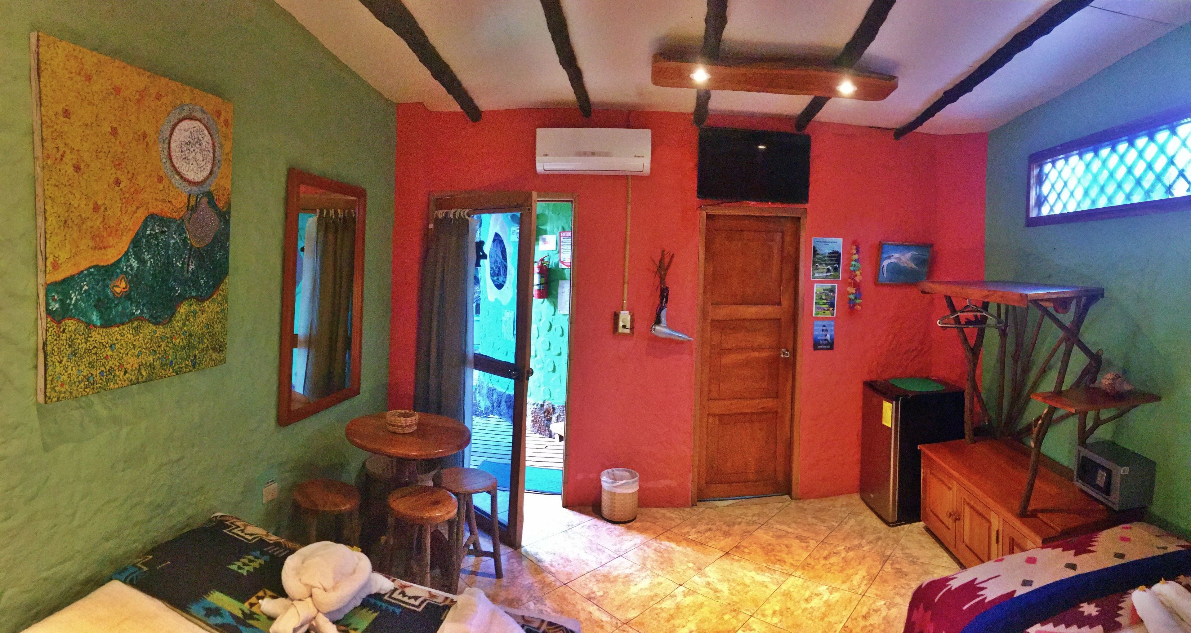 A room with a wooden ceiling, a door, a small table with stools, a bed, and a wall-mounted TV.