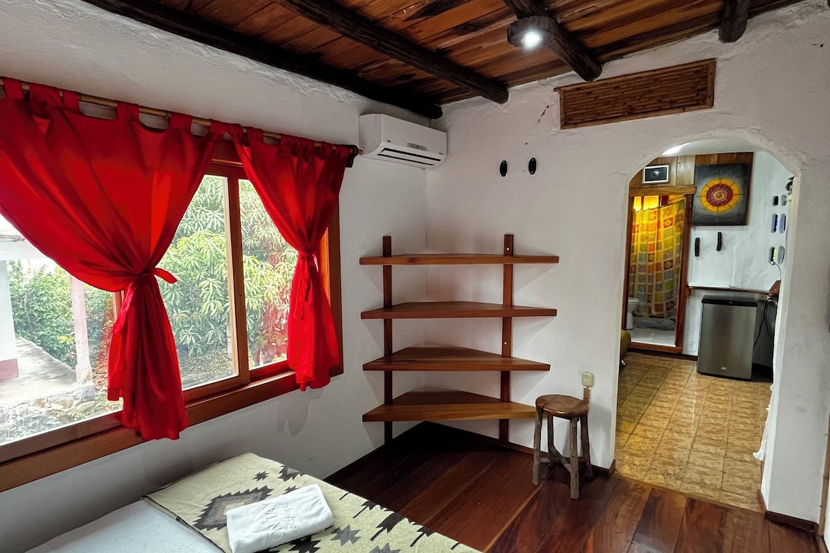 A room with a wooden ceiling, a bed with a patterned bedspread, a red curtain, and a small stool.