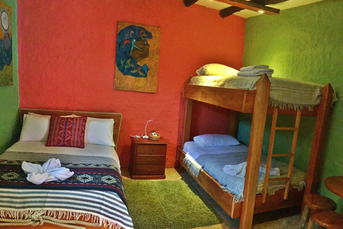 A room with a wooden bunk bed, a single bed with a patterned blanket, a small wooden nightstand, and a green carpet.