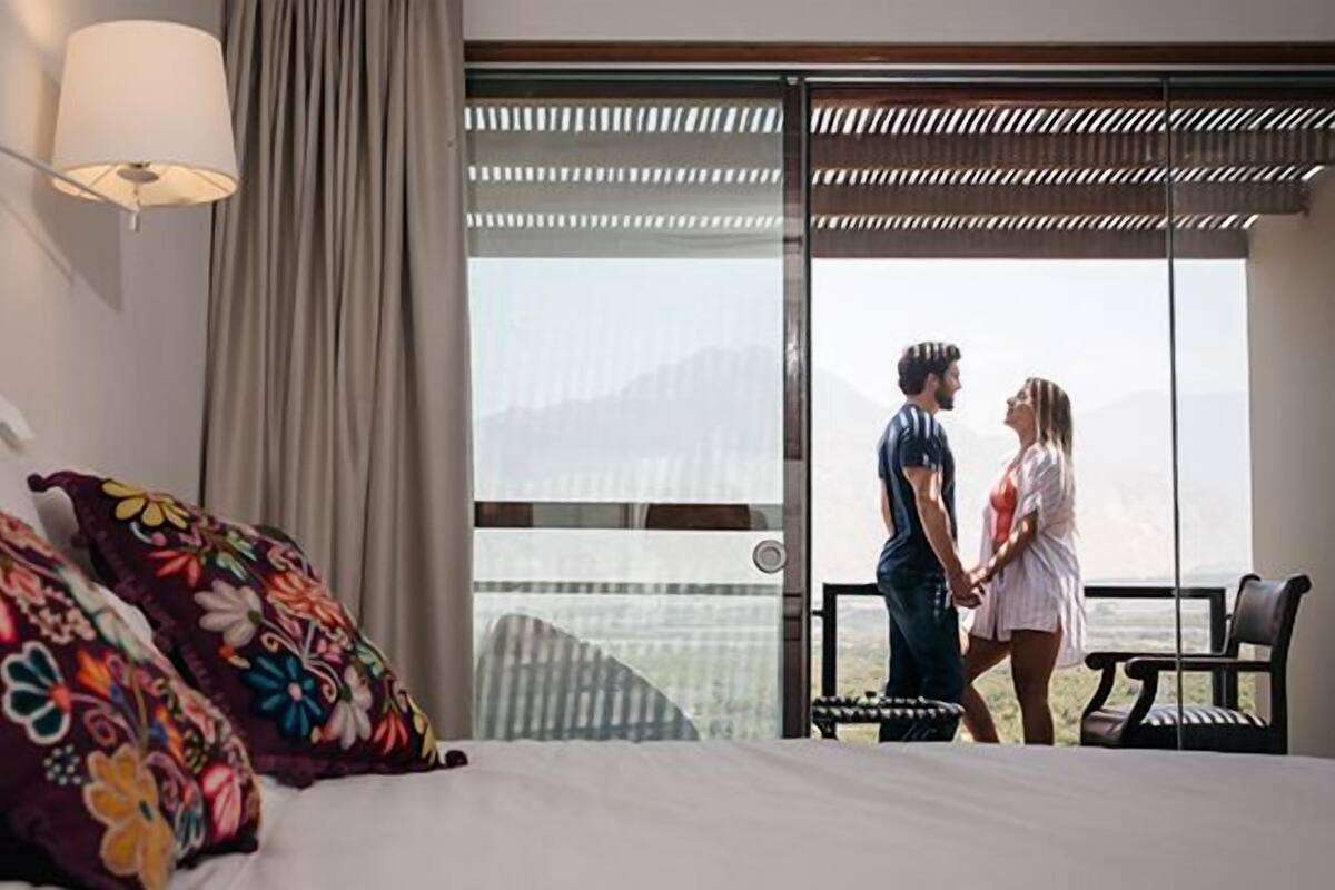 A hotel room with a bed, colorful pillows, a nightstand, a lamp, a large window with a view, and a balcony with a chair.