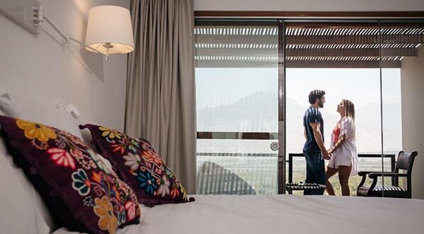 A hotel room with a bed, colorful pillows, a nightstand, a lamp, a large window with a view, and a balcony with a chair.