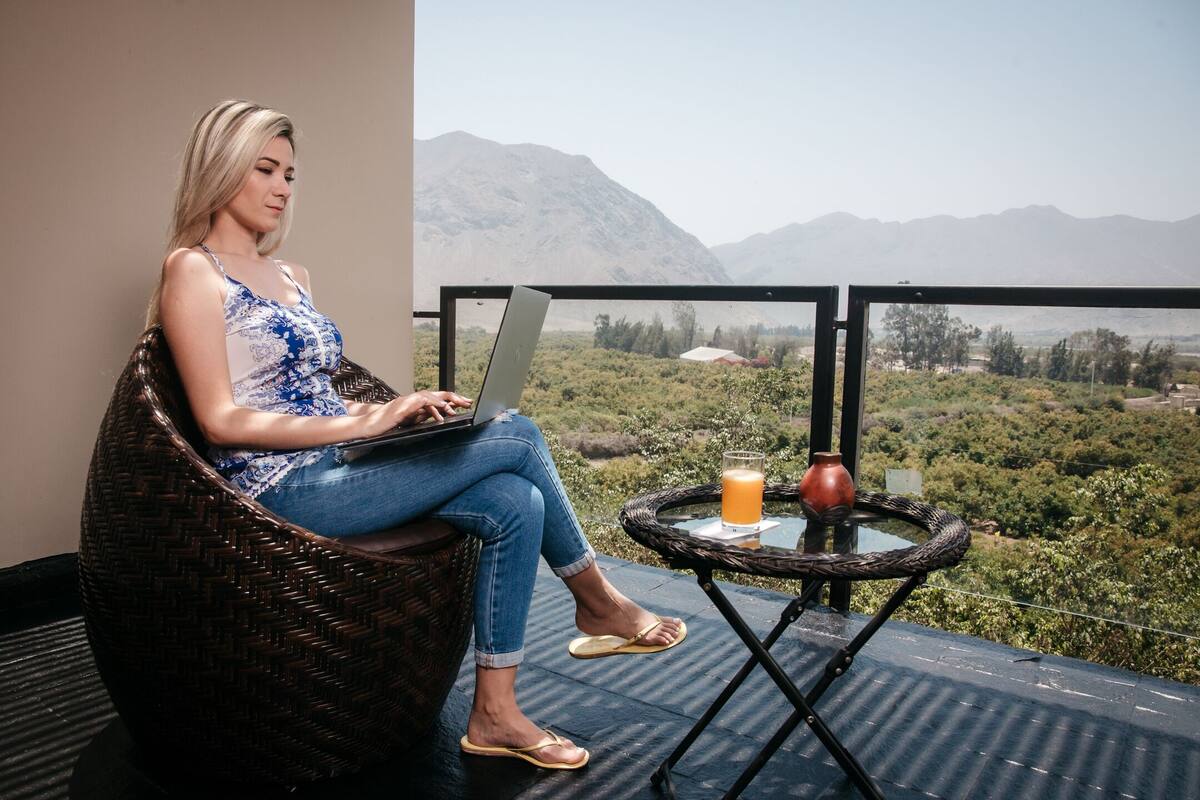 A person using a laptop on a wicker chair with a view of mountains and greenery.