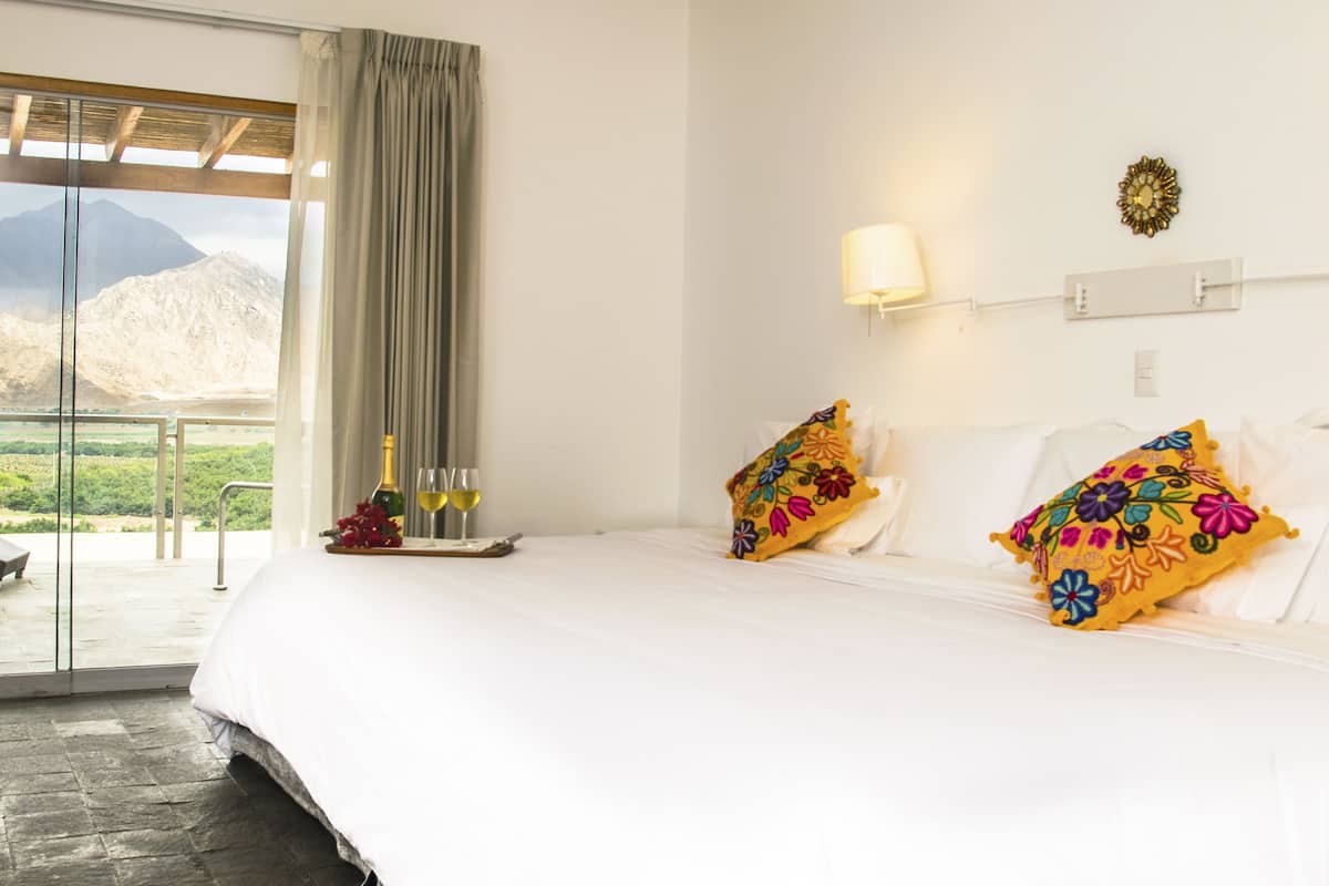 A hotel room with a bed, a nightstand with a bottle and glasses, a window with a view of mountains, and a balcony with a chair.