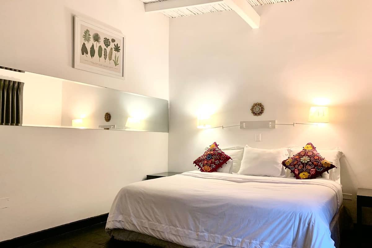 A neatly made bed with white bedding, two decorative pillows, a wall-mounted light fixture, and a framed botanical print above the bed.
