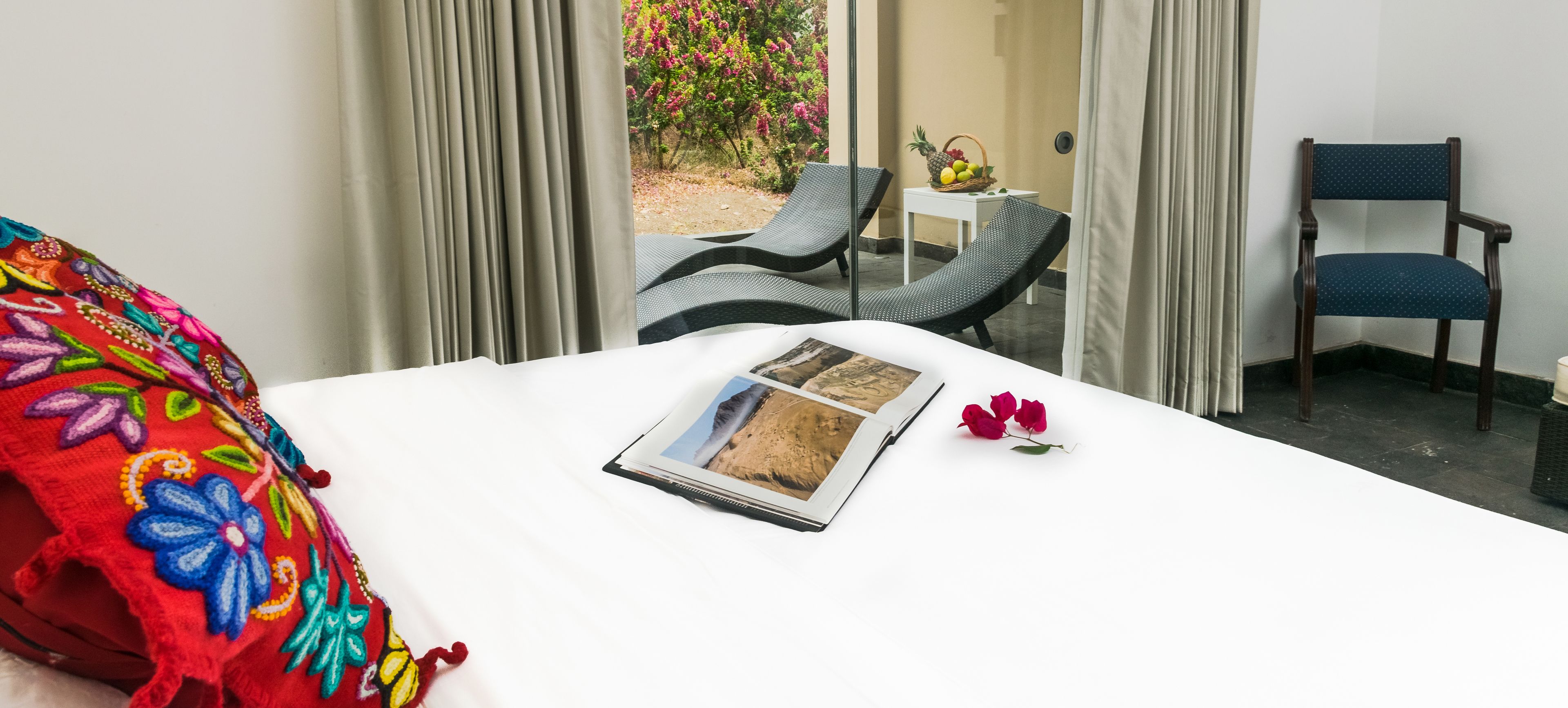 Une chambre d’hôtel avec un lit, une fenêtre avec des rideaux, une petite table sur laquelle se trouve un panier de fruits, et une chaise.