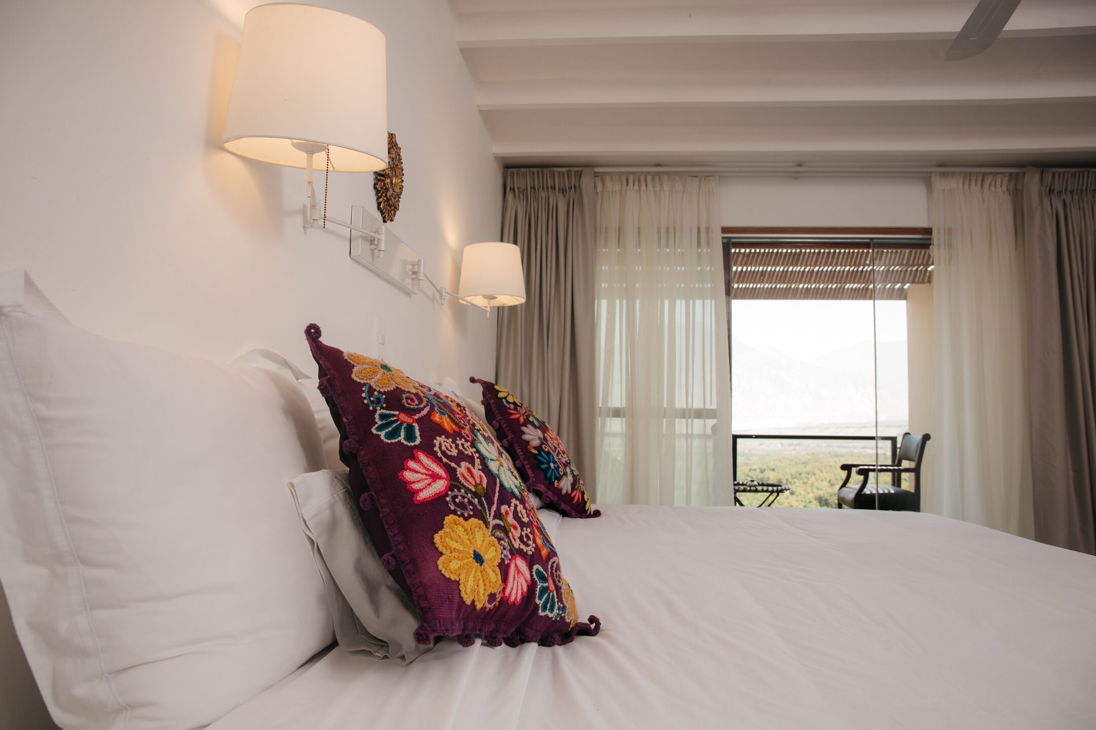 Habitación de hotel con una cama grande, dos almohadones decorativos, una mesita de noche y una ventana con cortinas.