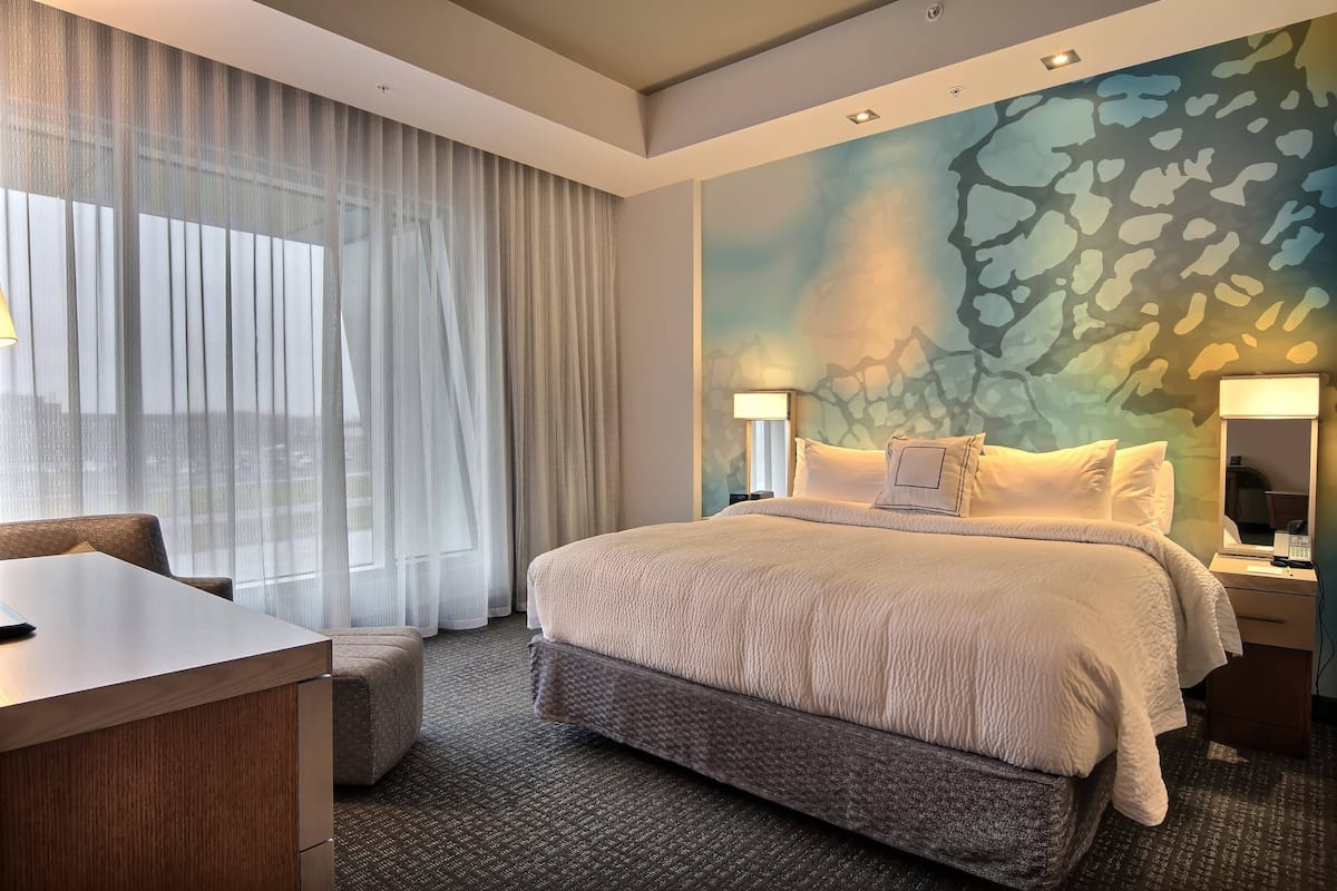 A hotel room with a large bed, a desk, and a chair. There is a window with sheer curtains and a wall with a textured mural.