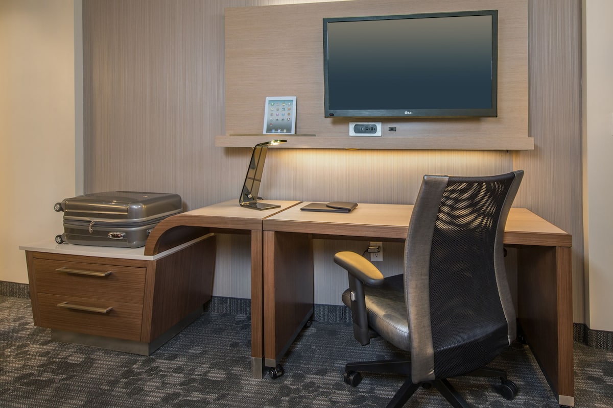 Habitación de hotel con un escritorio de madera, una silla de oficina negra, un televisor de pantalla plana en la pared, una lámpara de escritorio, una maleta y un pequeño panel de control.