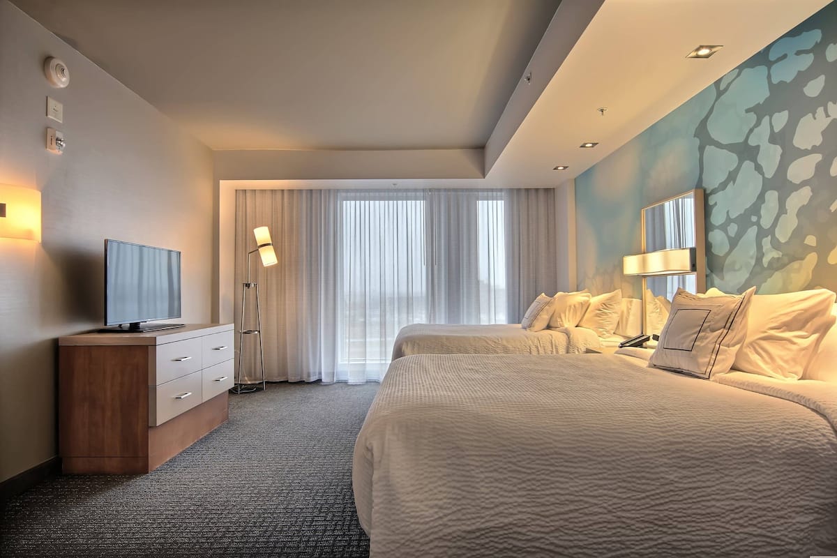 A hotel room with a large bed, a TV on a wooden cabinet, and a window with sheer curtains.