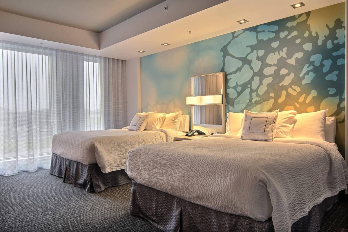 A hotel room with two beds, a large window with sheer curtains, a wall with a textured mural, and a ceiling with recessed lighting.