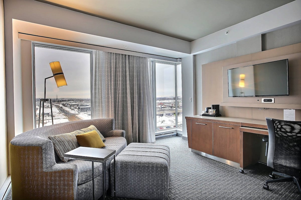 A modern hotel room with a large window, a sofa, a desk, and a TV.