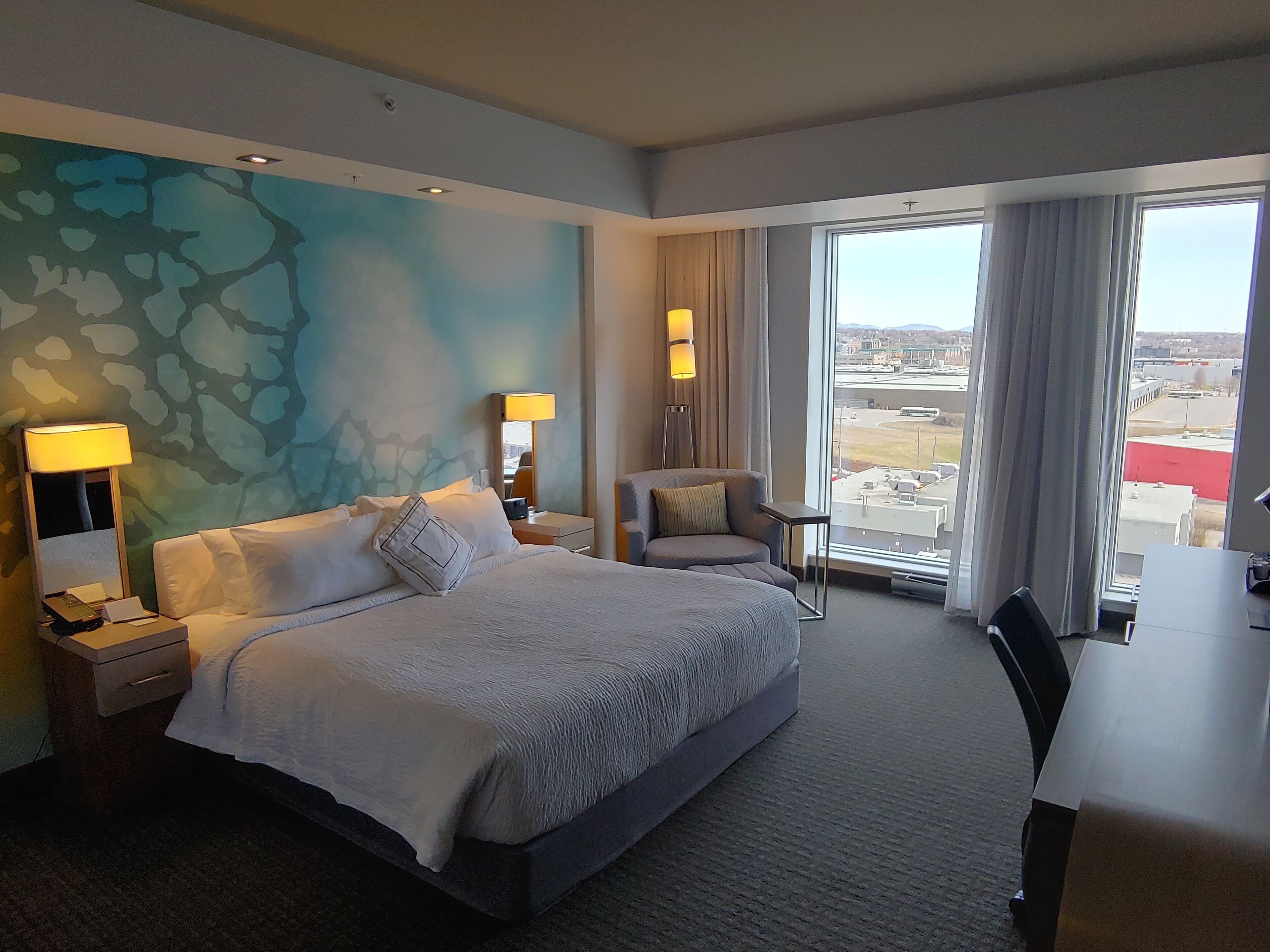 A hotel room with a large bed, a desk, a chair, and a view of the airport.
