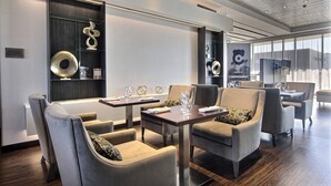 Un moderno vestíbulo de hotel con comedor que incluye sofás y mesas, un ventanal con cortinas y un televisor montado en la pared.