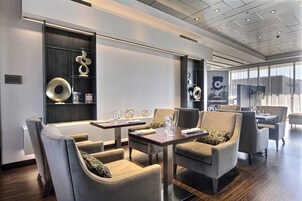 Un moderno vestíbulo de hotel con comedor que incluye sofás y mesas, un ventanal con cortinas y un televisor montado en la pared.