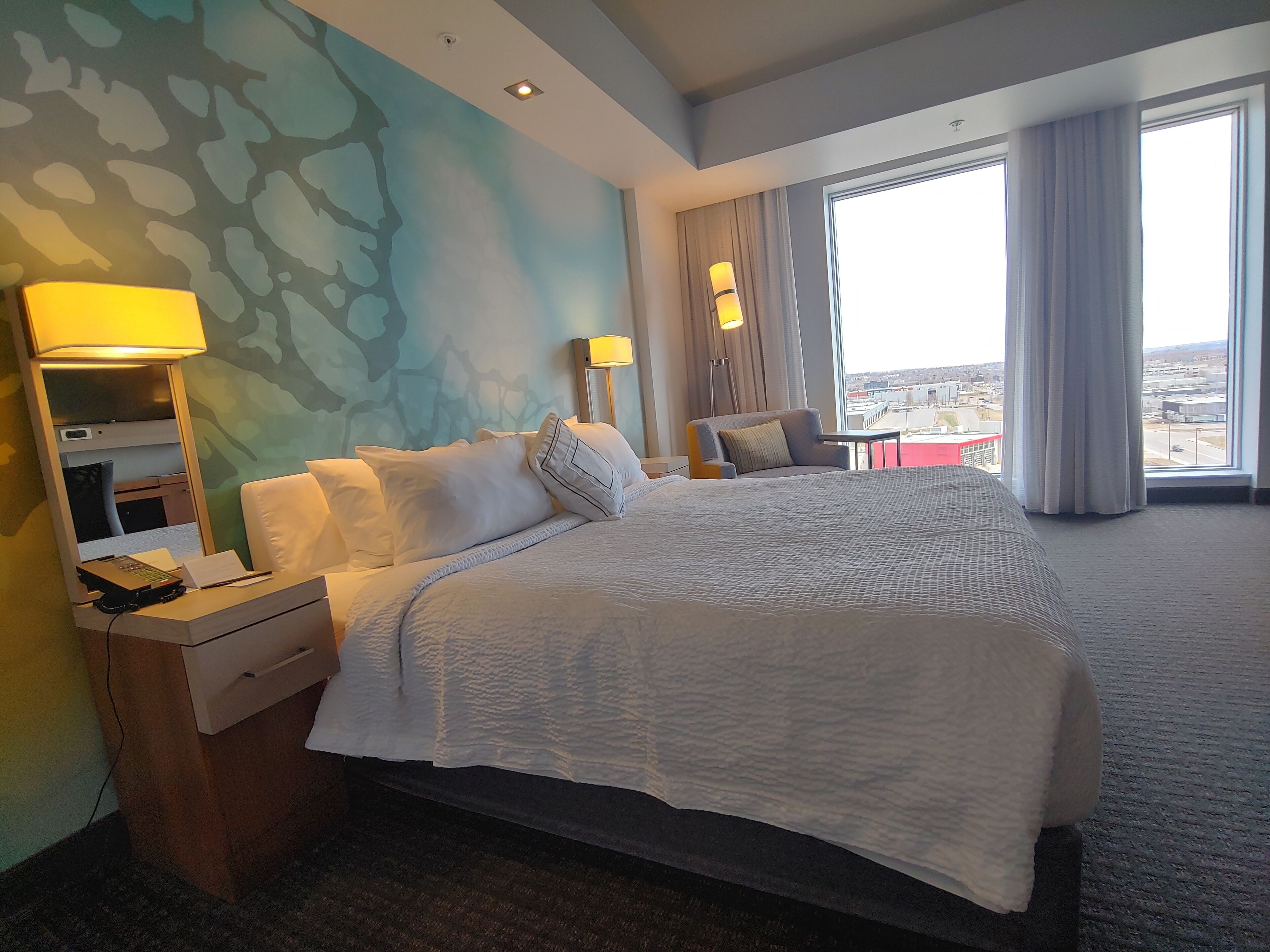 A hotel room with a large bed, bedside tables, a desk, and a view of the city through large windows.