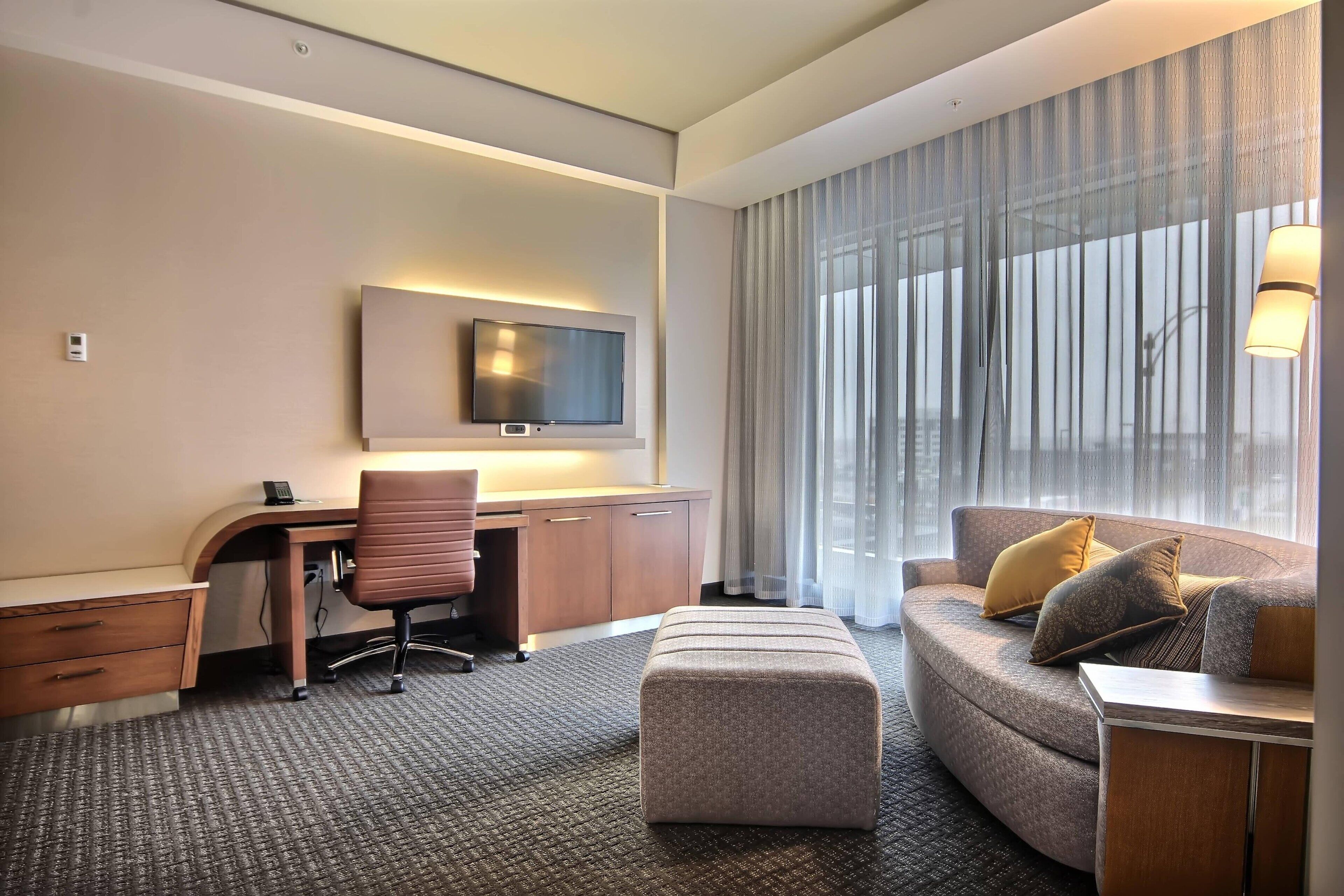 Una habitación de hotel moderna con un sofá curvo, un escritorio con una silla, un televisor montado en la pared y un ventanal con cortinas translúcidas.