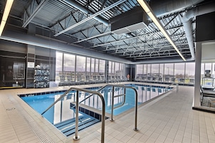 Una piscina interior con agua cristalina, barandales de metal y vistas a la ciudad a través de amplias ventanas.