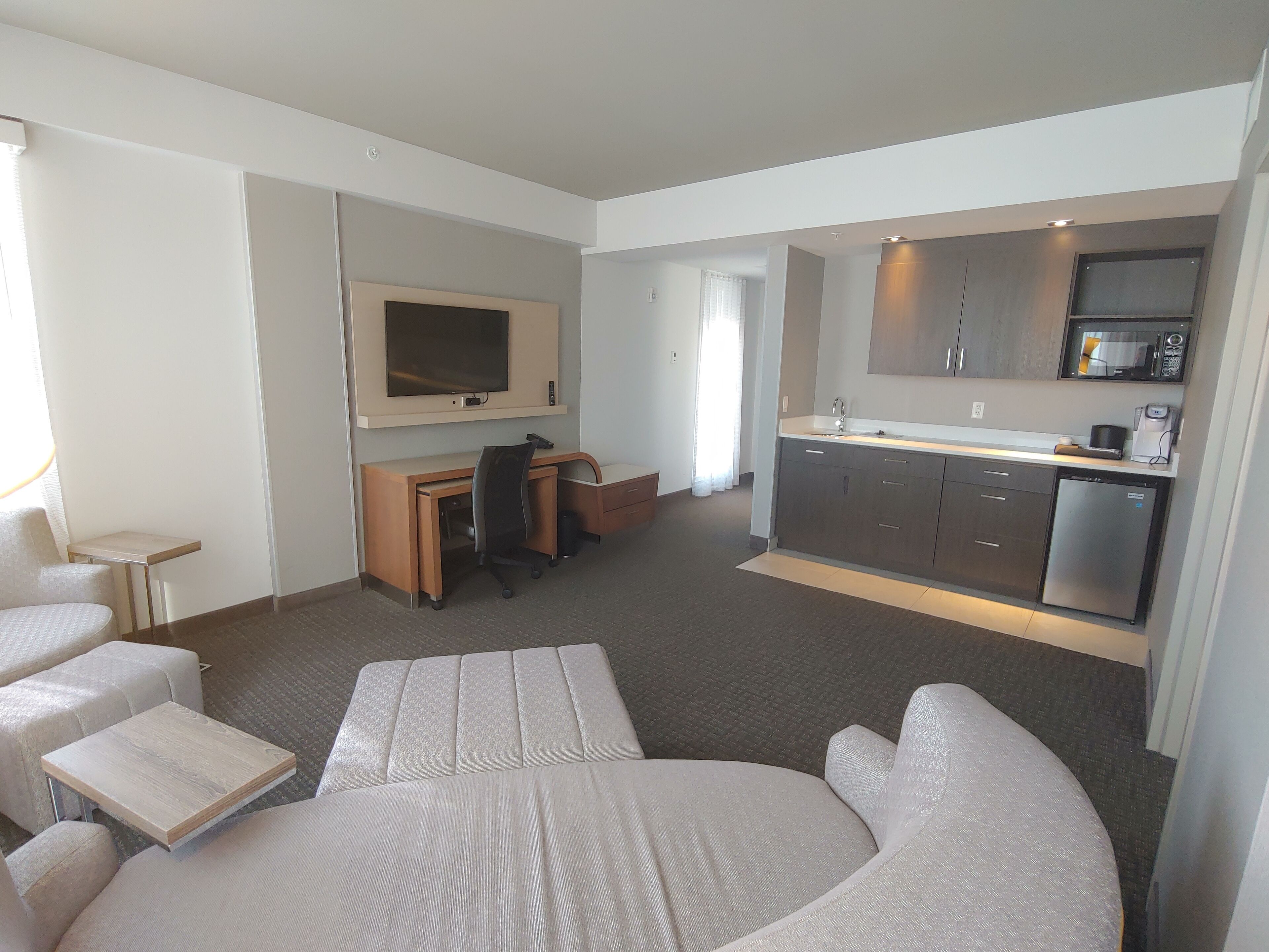 A modern hotel room with a bed, a desk, a TV, a kitchenette, and a seating area.