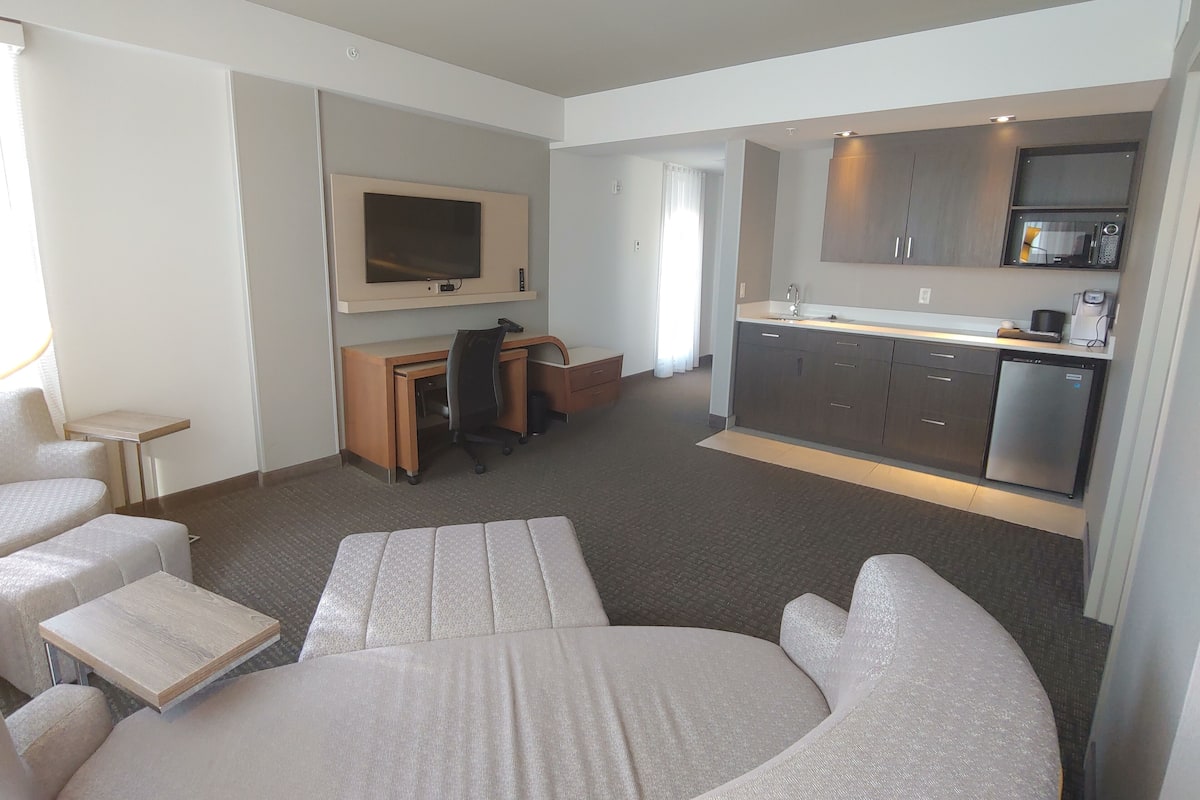 Una habitación de hotel moderna con cama, escritorio, televisor, kitchenette y zona de estar.
