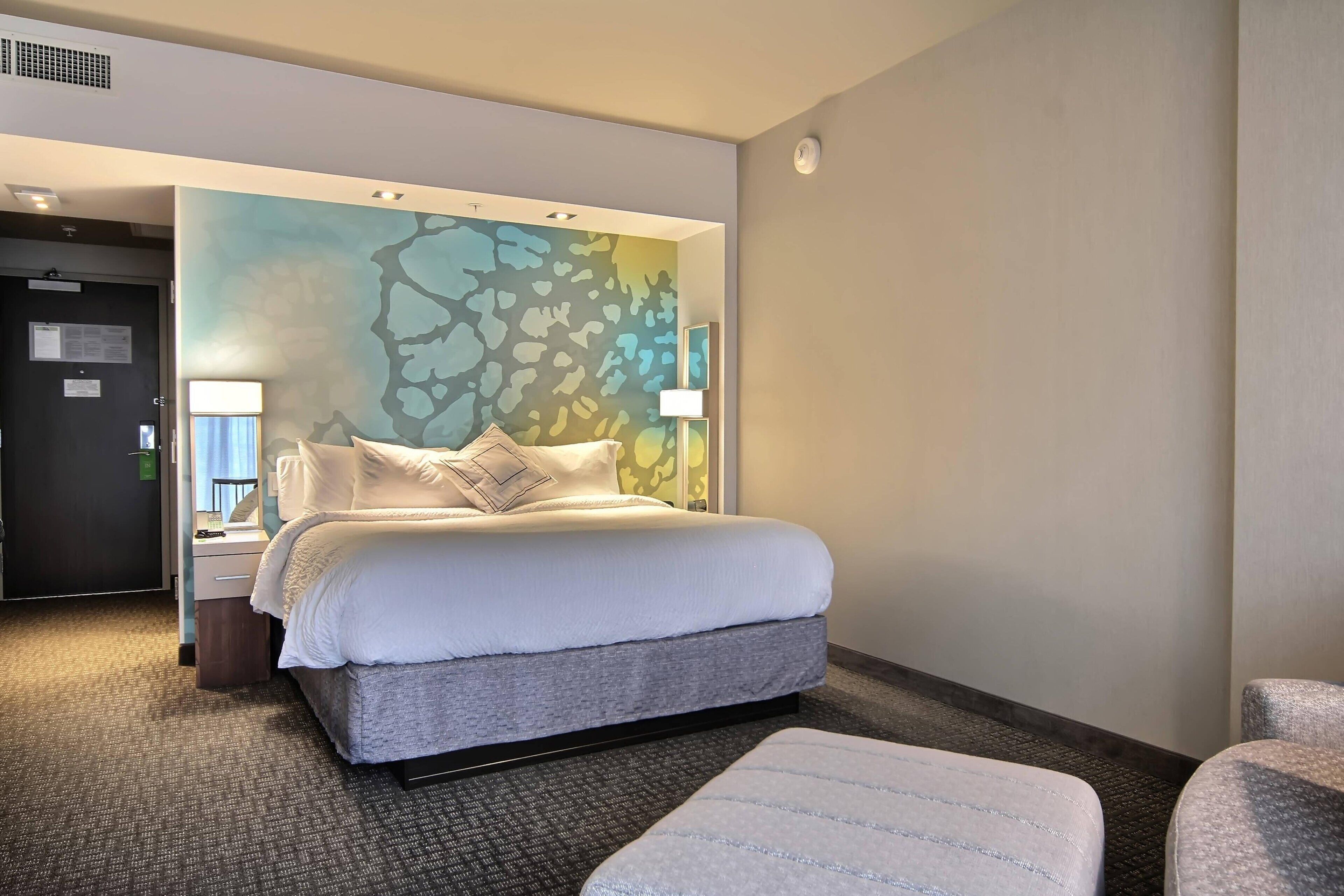 A hotel room with a bed, a nightstand, a lamp, a chair, and a carpeted floor.