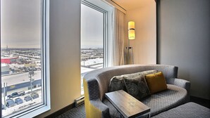 Una habitación de hotel moderna con un sofá gris curvo, una pequeña mesita de madera y una lámpara de pie. Un ventanal grande ofrece vistas de la ciudad.