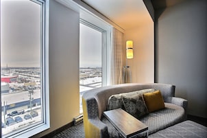 Una habitación de hotel moderna con un sofá gris curvo, una pequeña mesita de madera y una lámpara de pie. Un ventanal grande ofrece vistas de la ciudad.