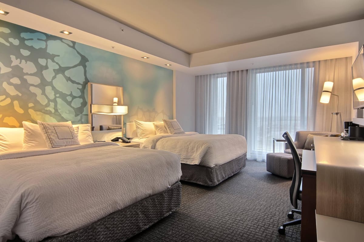 A hotel room with two beds, a desk, a chair, and a large window with curtains.