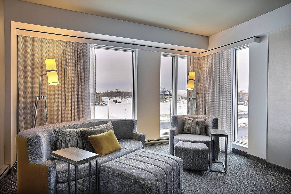 A modern hotel room with a grey sofa, a small table, and a chair. There are large windows with curtains and two wall-mounted lamps.