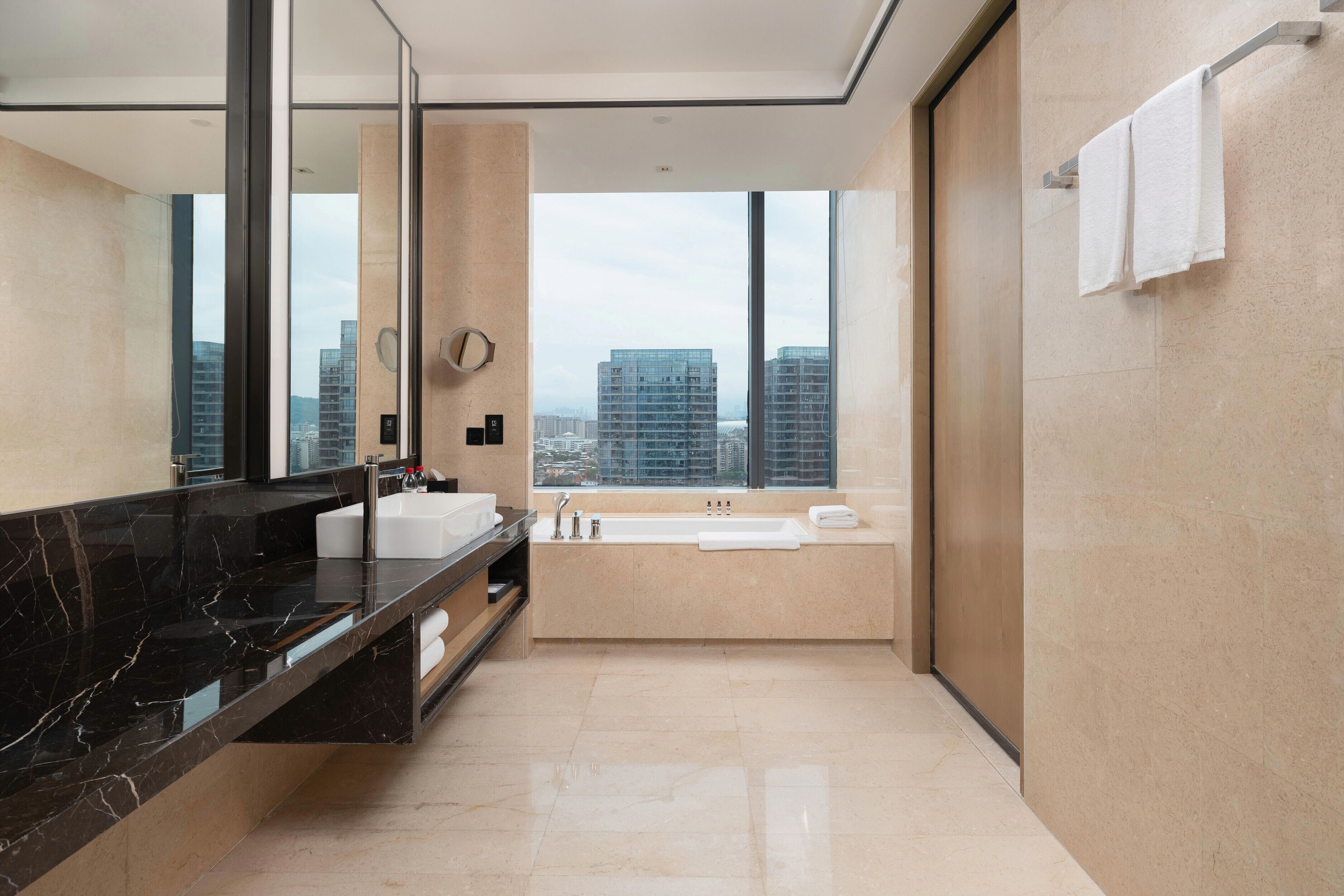 A modern bathroom with a large mirror, a black marble countertop, a bathtub, and a view of tall buildings.