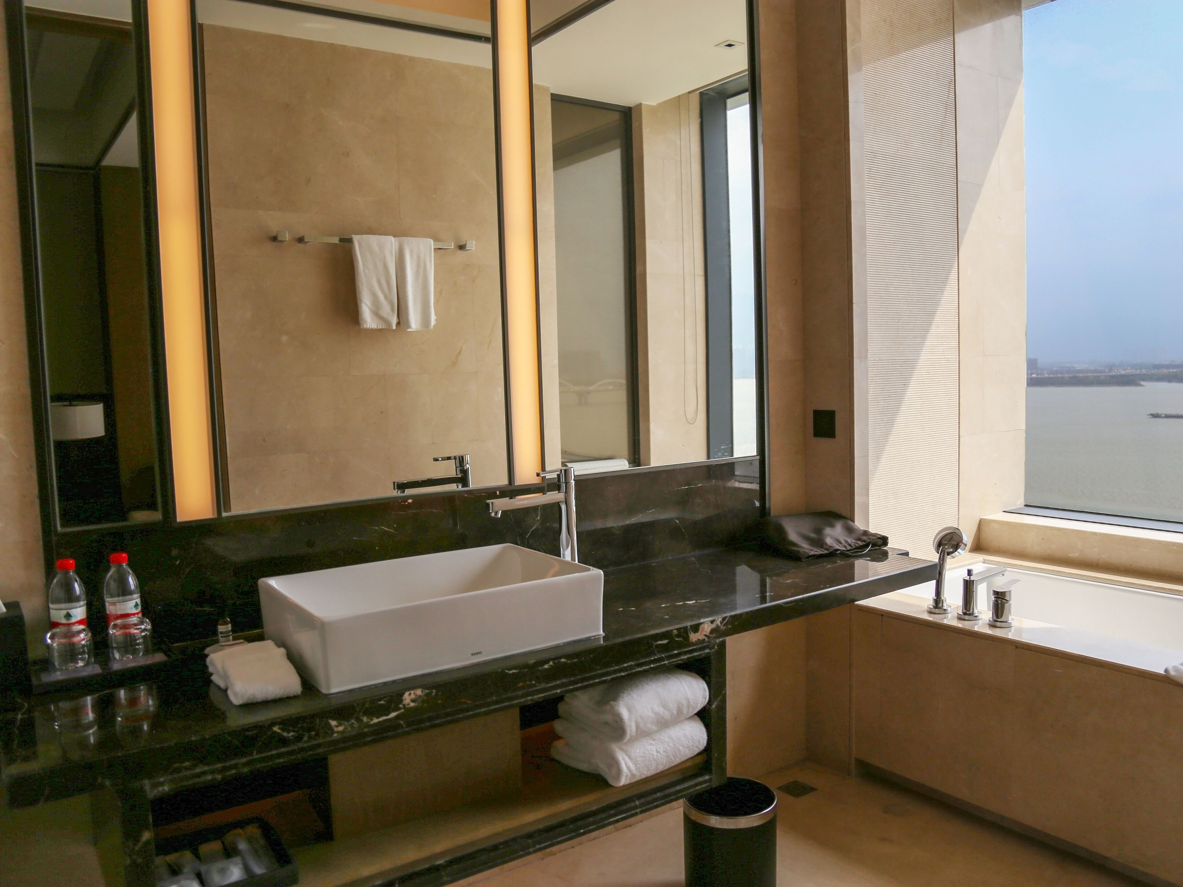 A modern bathroom with a large mirror, a white sink, and a bathtub. There are towels and bottles on the counter.