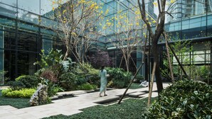 Un edificio moderno con fachadas de vidrio, un patio ajardinado con diversas plantas y una persona caminando.