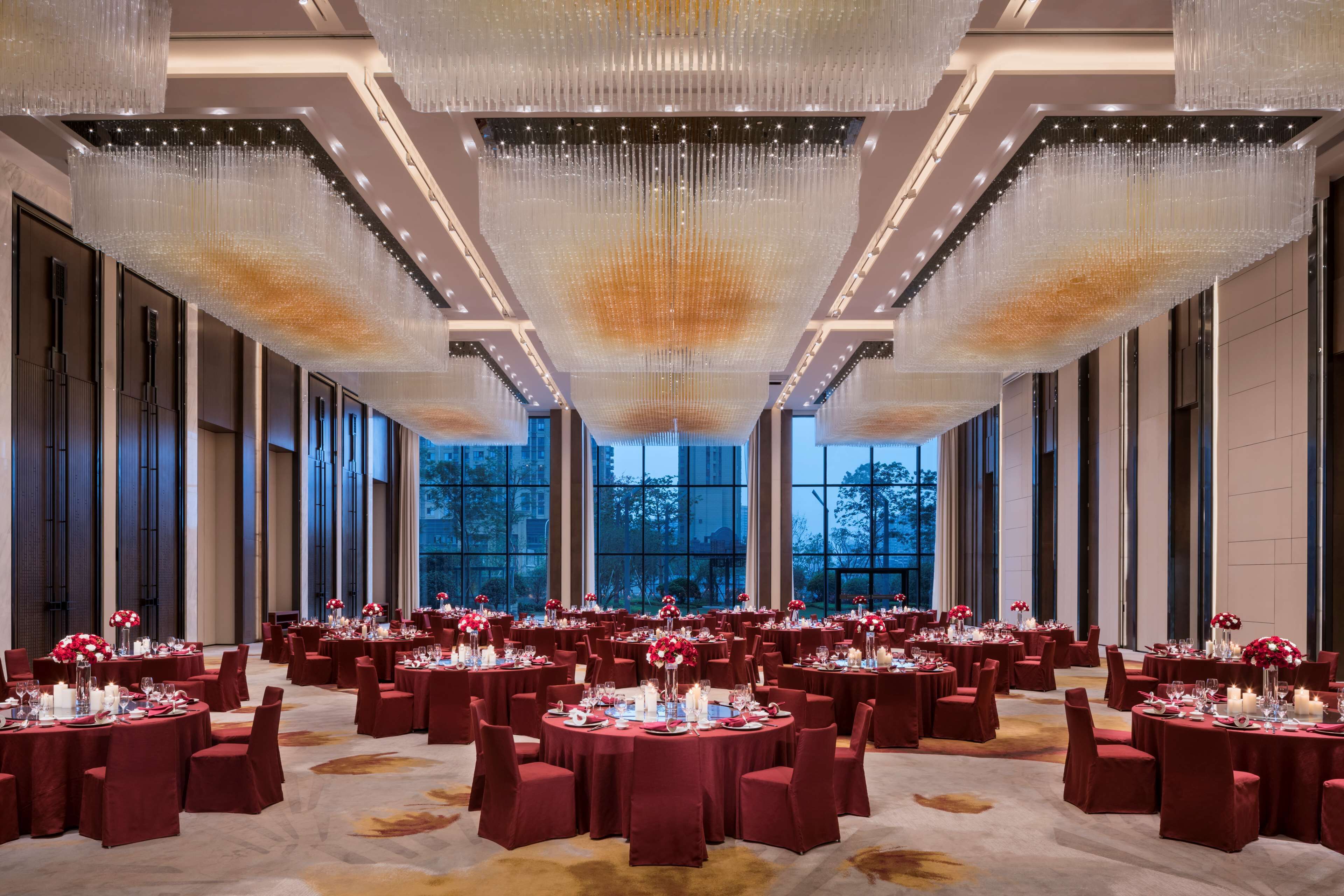 A spacious banquet hall with round tables set for dining, large windows, and a modern chandelier.
