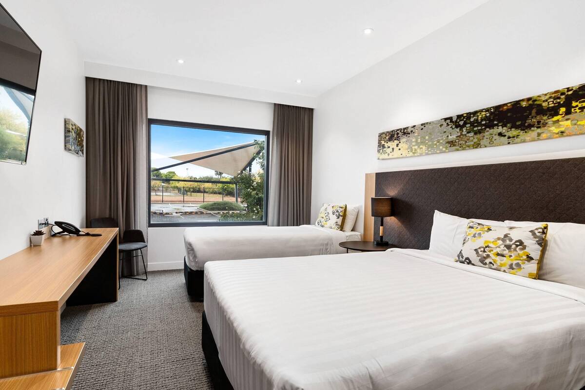 Een hotelkamer met twee bedden, een bureau, een stoel, een lamp en een groot raam met uitzicht op een gebouw en groen.