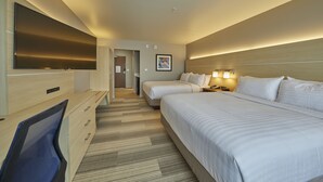 A hotel room with a large flat-screen TV, two beds, a desk with a chair, and a headboard with built-in lighting.