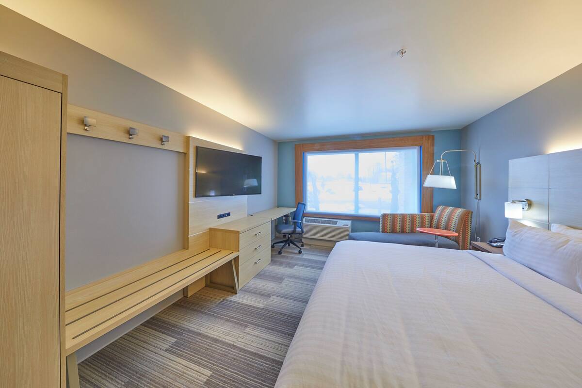 A hotel room with a large bed, a desk with a chair, a TV, a window, and a lamp.