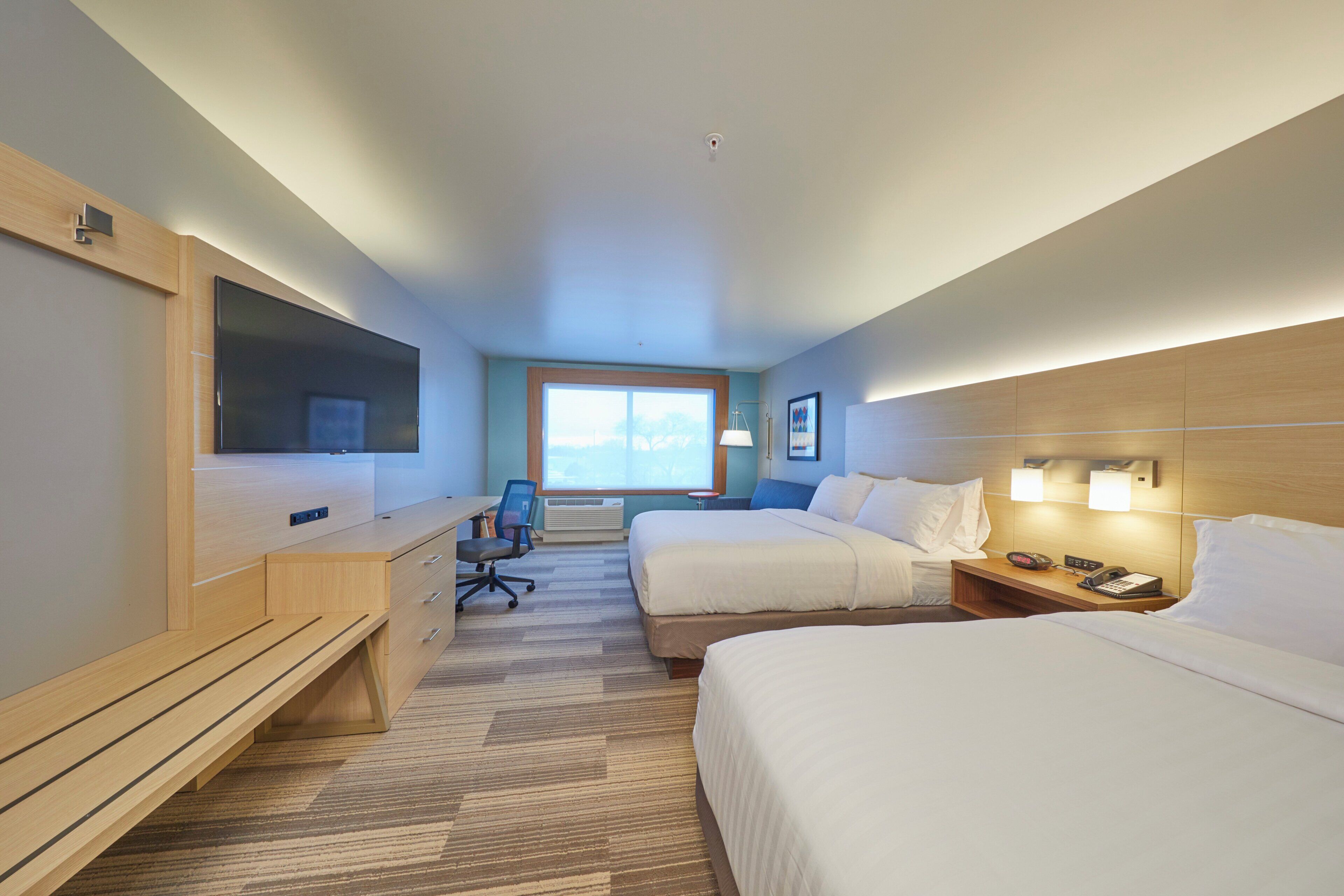 A hotel room with two beds, a desk, a chair, a TV, and a window.