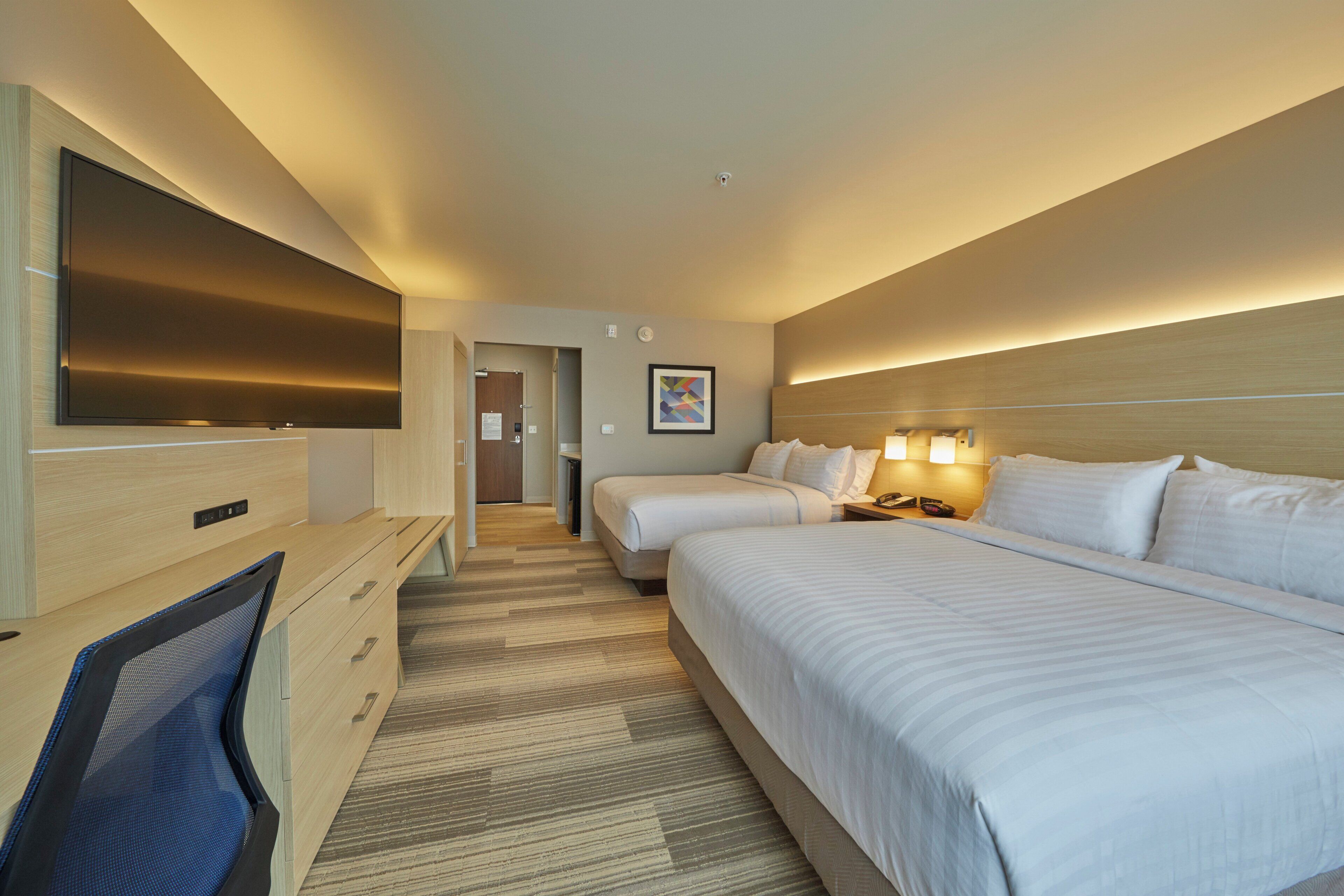 A hotel room with two beds, a desk, a flat-screen TV, and a headboard with integrated lighting.